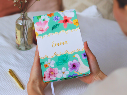 Person holding a notebook with floral design, gold foiled name and personalised custom name next on a white surface.