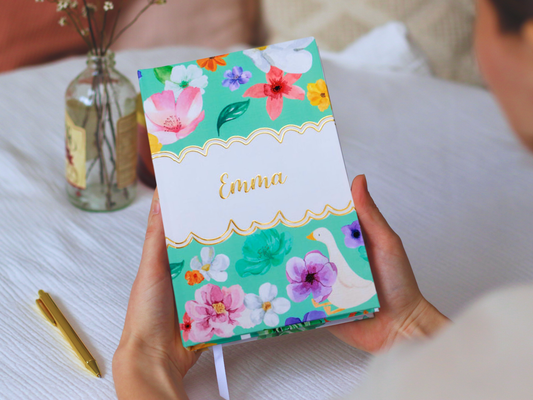 Person holding a notebook with floral design, gold foiled name and personalised custom name next on a white surface.